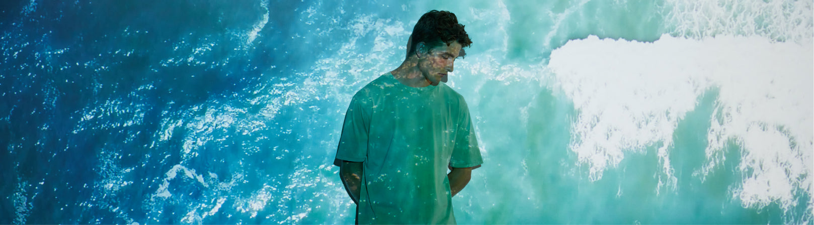 Person in a stone washed green shirt standing in front of a large wave, plastic free no microplastic no polyester apparel earth positive t-shirt EP185.